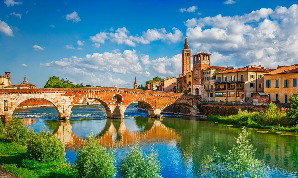 Enjoying a Parodi cigar and a panoramic view to Bridge Ponte Pietra in Verona on Adige river, Veneto region, Italy. Sunny summer day panorama and blue dramatic sky with clouds. Ancient european italian terracotta color houses.